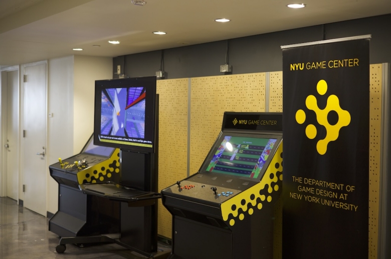 Photo of two vintage arcade games at the NYU Game Center. To the right is a standing black banner with yellow text that reads NYU GAME CENTER THE DEPARTMENT OF GAME DESIGN AT NEW YORK UNIVERSITY in all caps